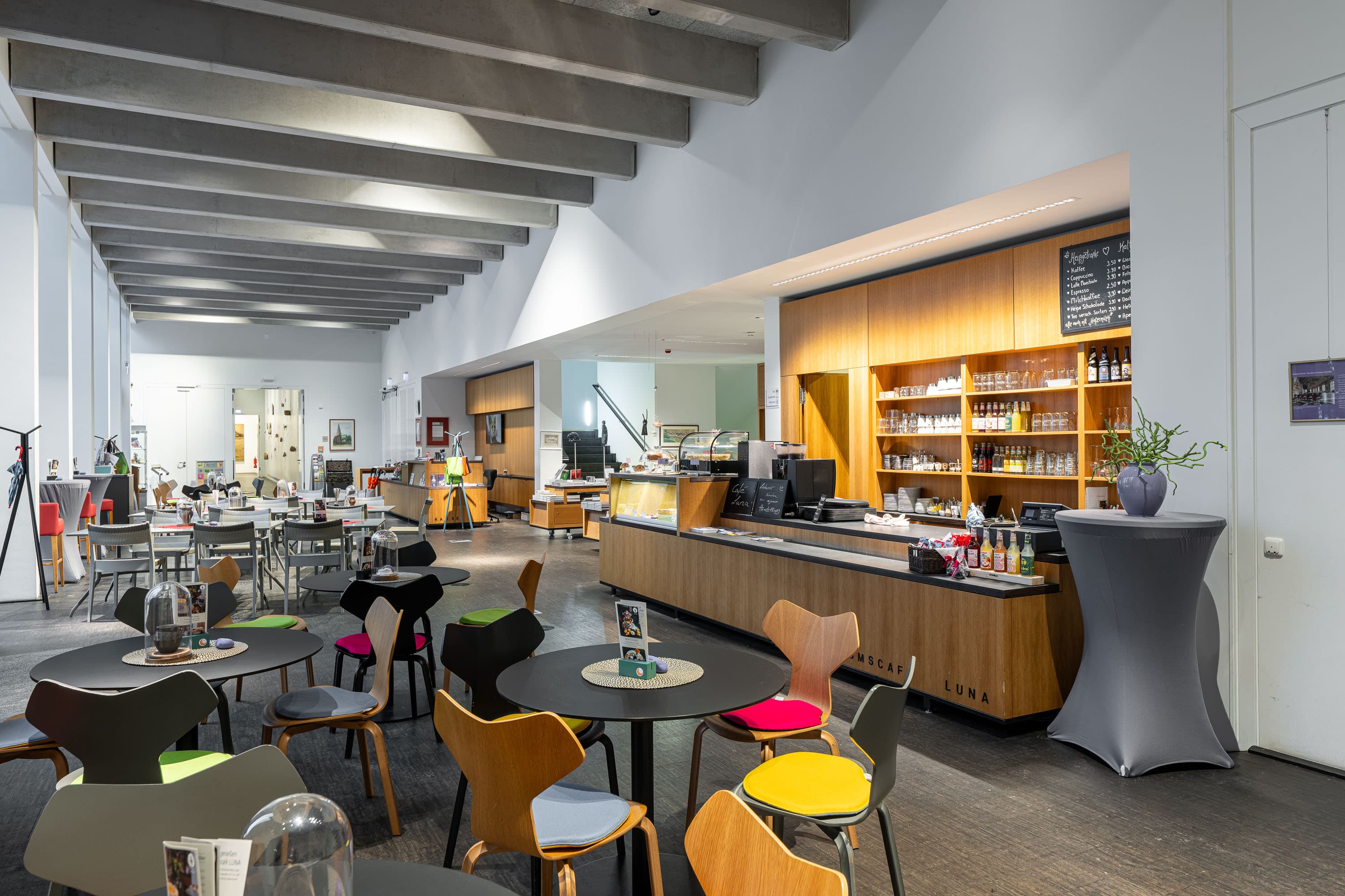 Heller Innenraum des Museumscafés LUNA mit modernem Tresen und bunten Stühlen an runden Tischen.