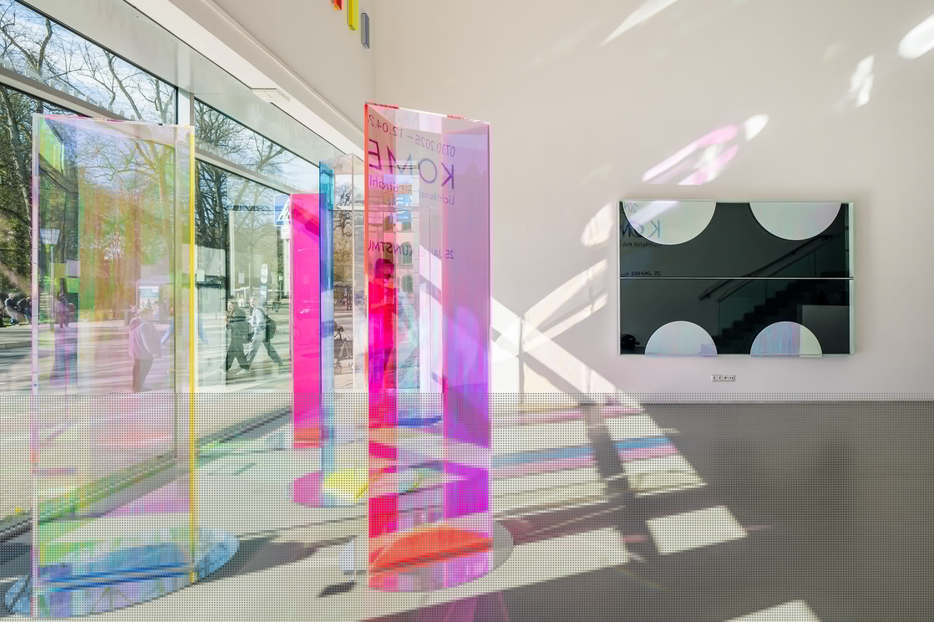 Lichtdurchfluteter Raum des Eingangsbereichs mit bunten, transparenten Stelen, die farbige Schatten werfen. Kunst von Regine Schumann