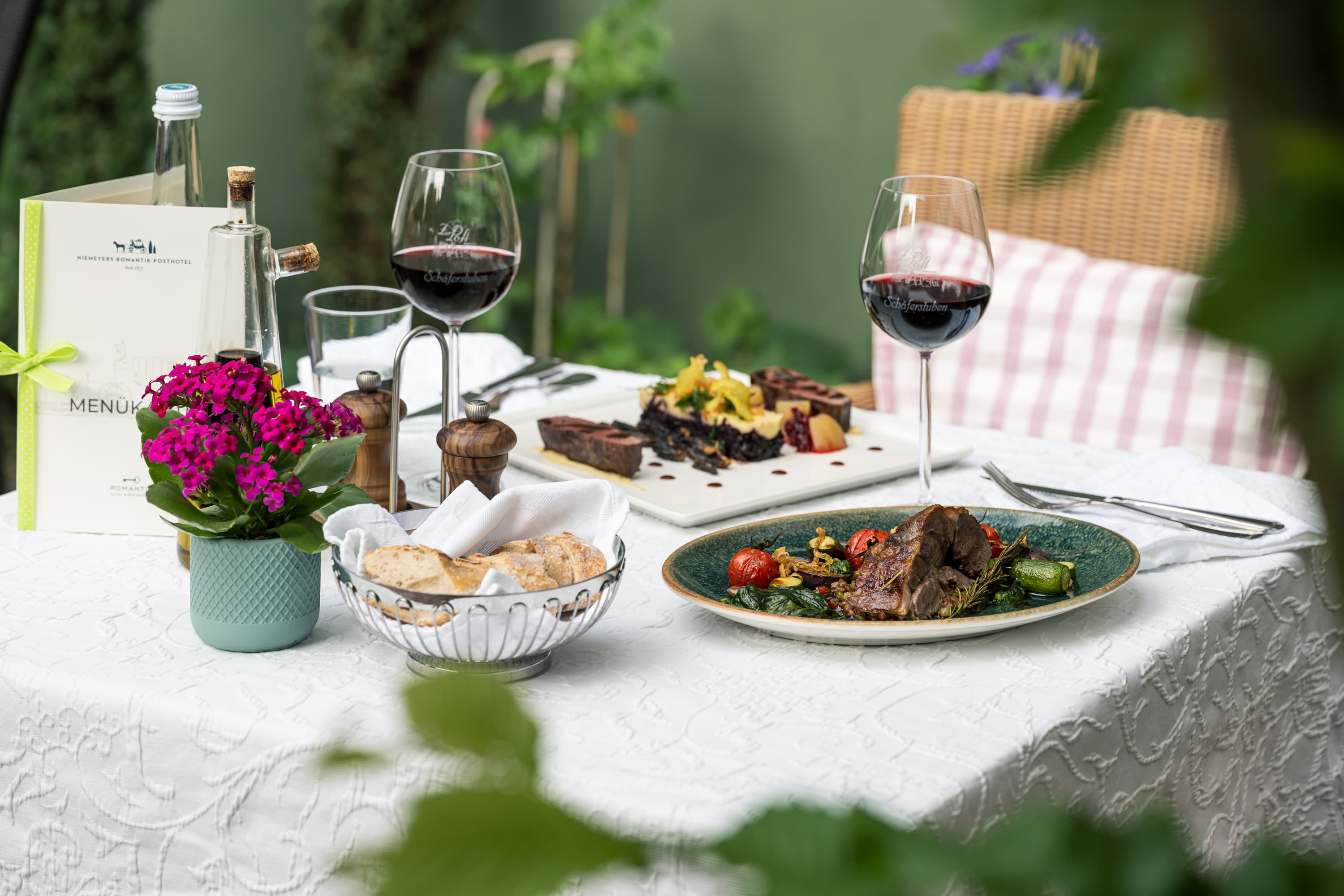 Zwei Teller mit Heidschnucken-Spezialitäten und Rotwein auf einem weiß gedeckten Tisch im Freien.