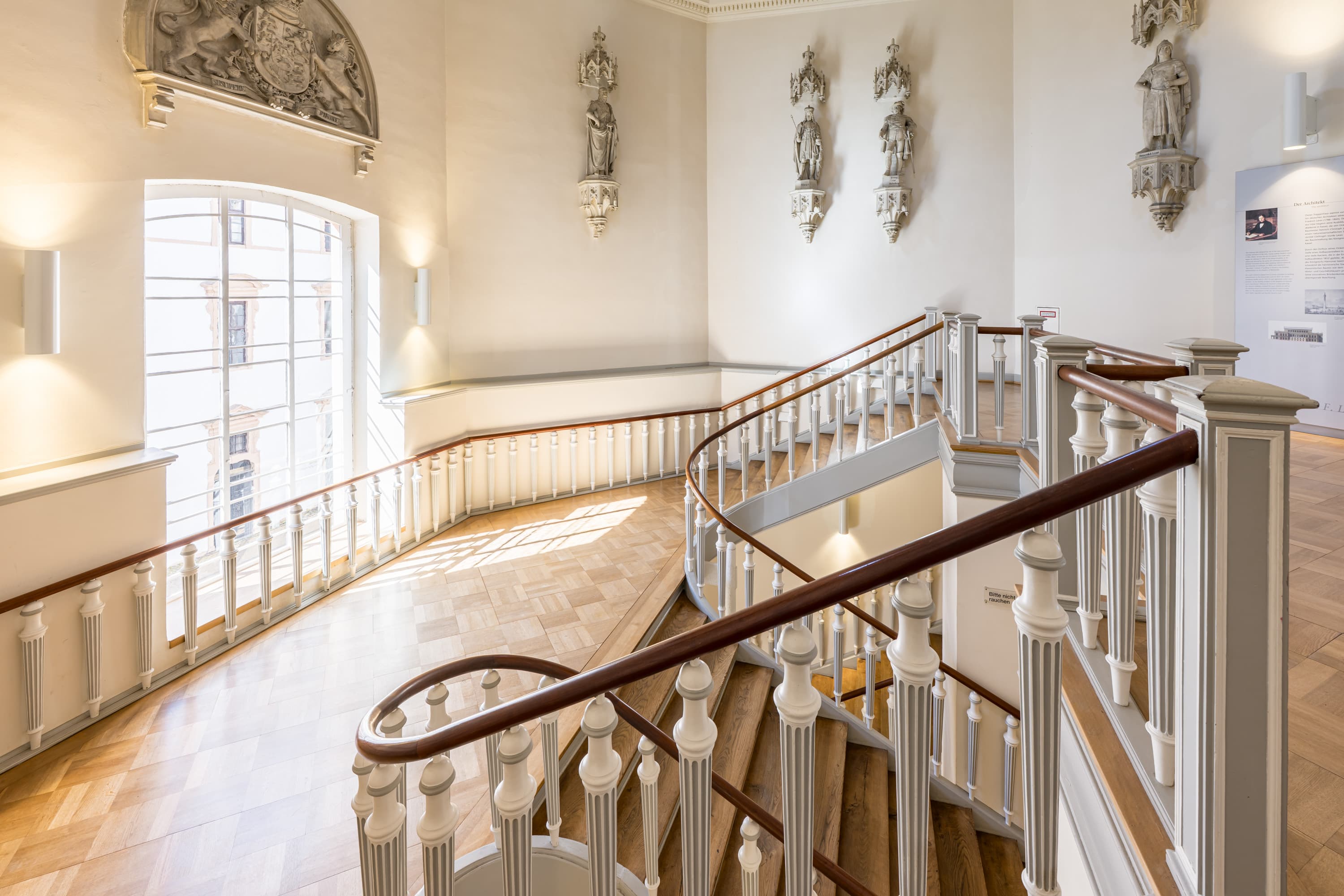 Das helle, geschwungene Treppenhaus im Celler Schloss mit verziertem Geländer und Parkettboden.