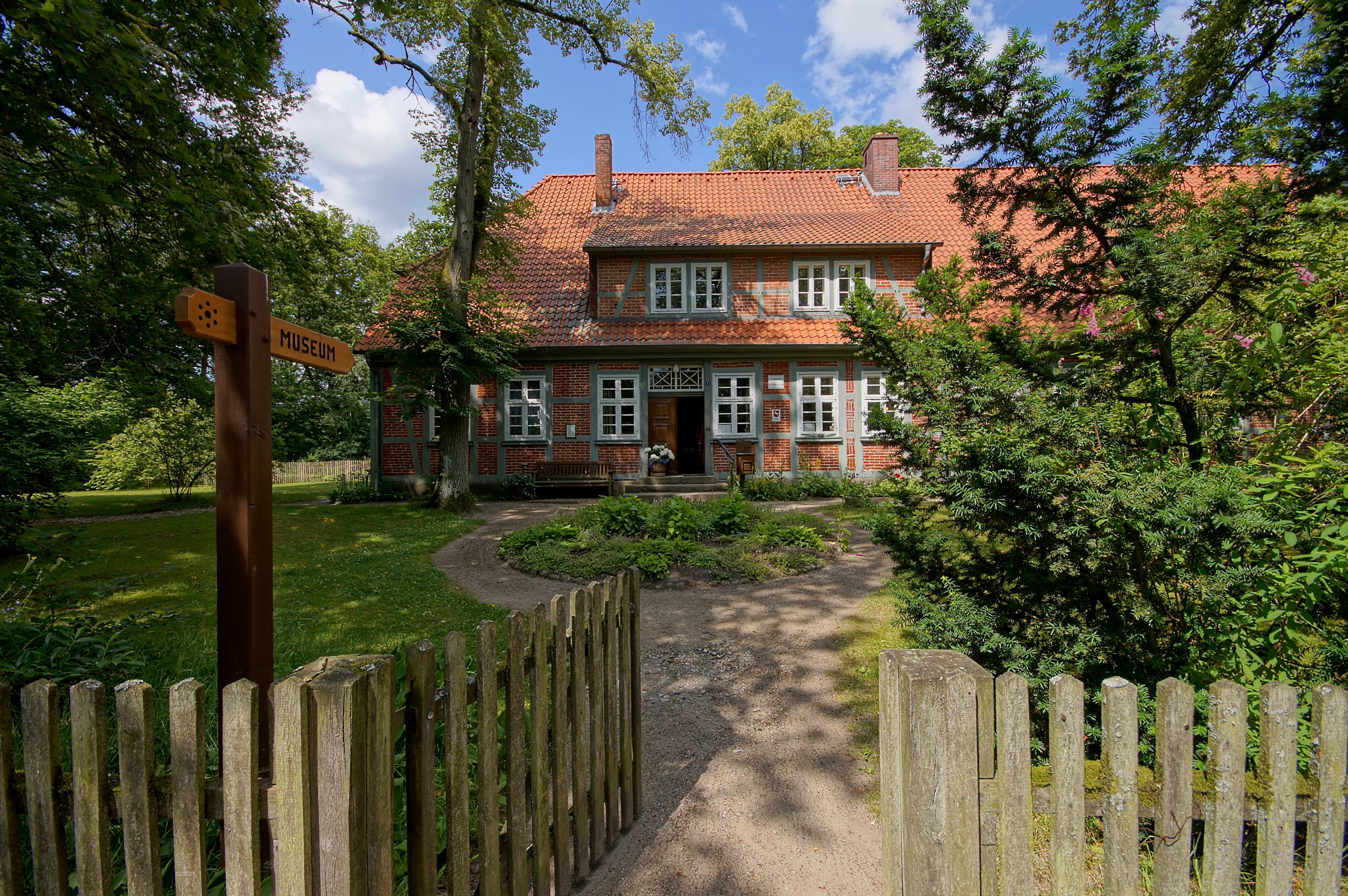 Ein traditionelles Backsteingebäude mit roten Dachziegeln, umgeben von üppigem Grün und Bäumen.A pathway leads to a red-brick building with white windows, surrounded by trees and greenery.